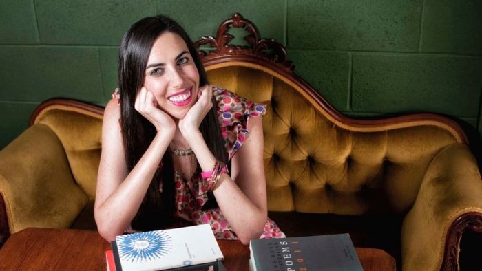 Woman sitting on a velvet couch leaning on a pile of books, smiling with long brown hair