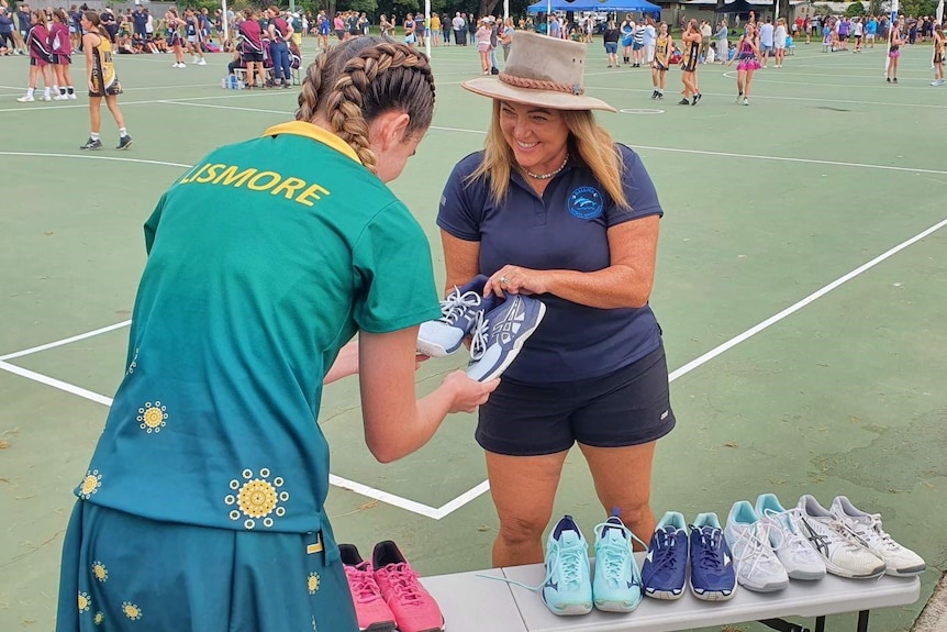 A woman stands in front of a bench with pairs of netball shoes. She hands over one pair to a girl wearing a Lismore uniform.