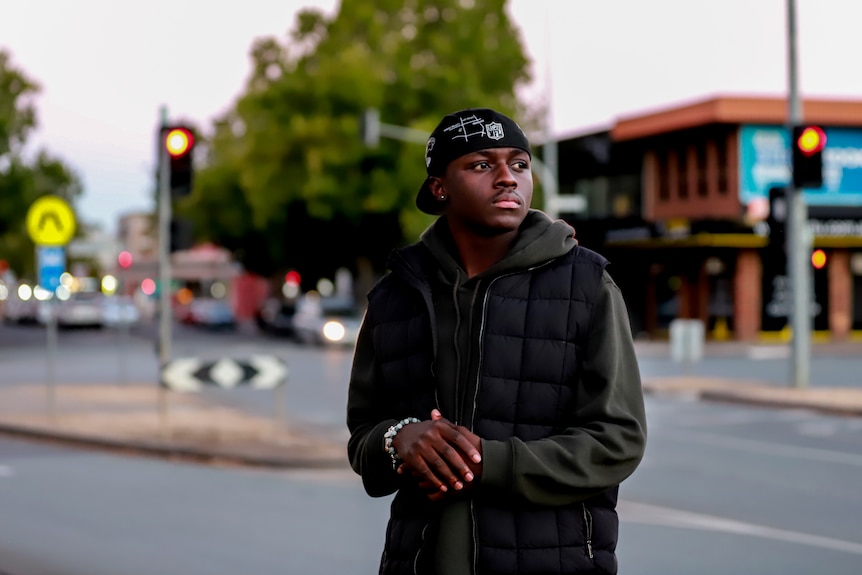 Man with dark skin stands in regional city street wearing black cap and puffer jacket