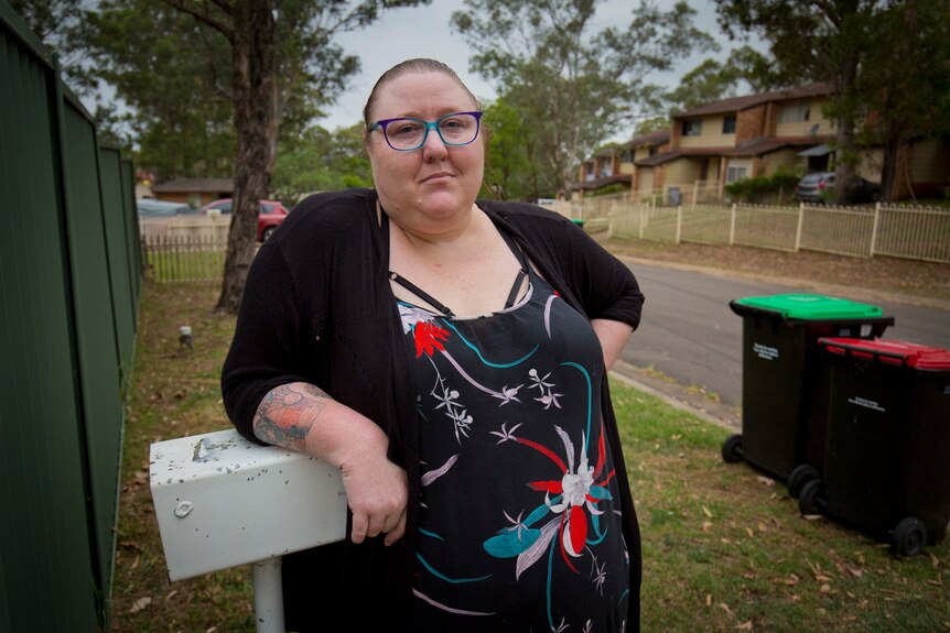 Sharmy Walker leans against a white mailbox next to a green fence. A suburban street and council rubbish bins are behind her.