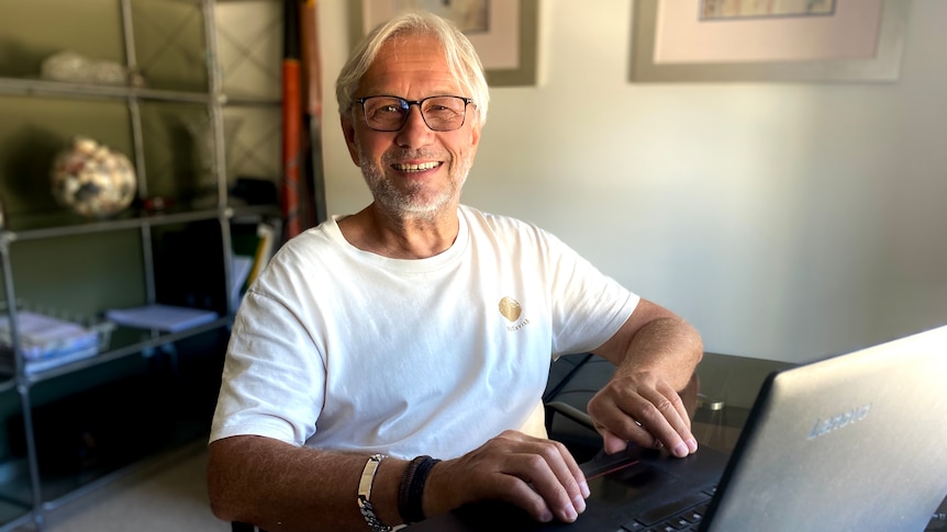 An older man in a white T-shirt smiles while typing on a computer.