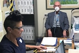 A bald man sits with a face mask on while a woman in scrubs stares at a laptop screen