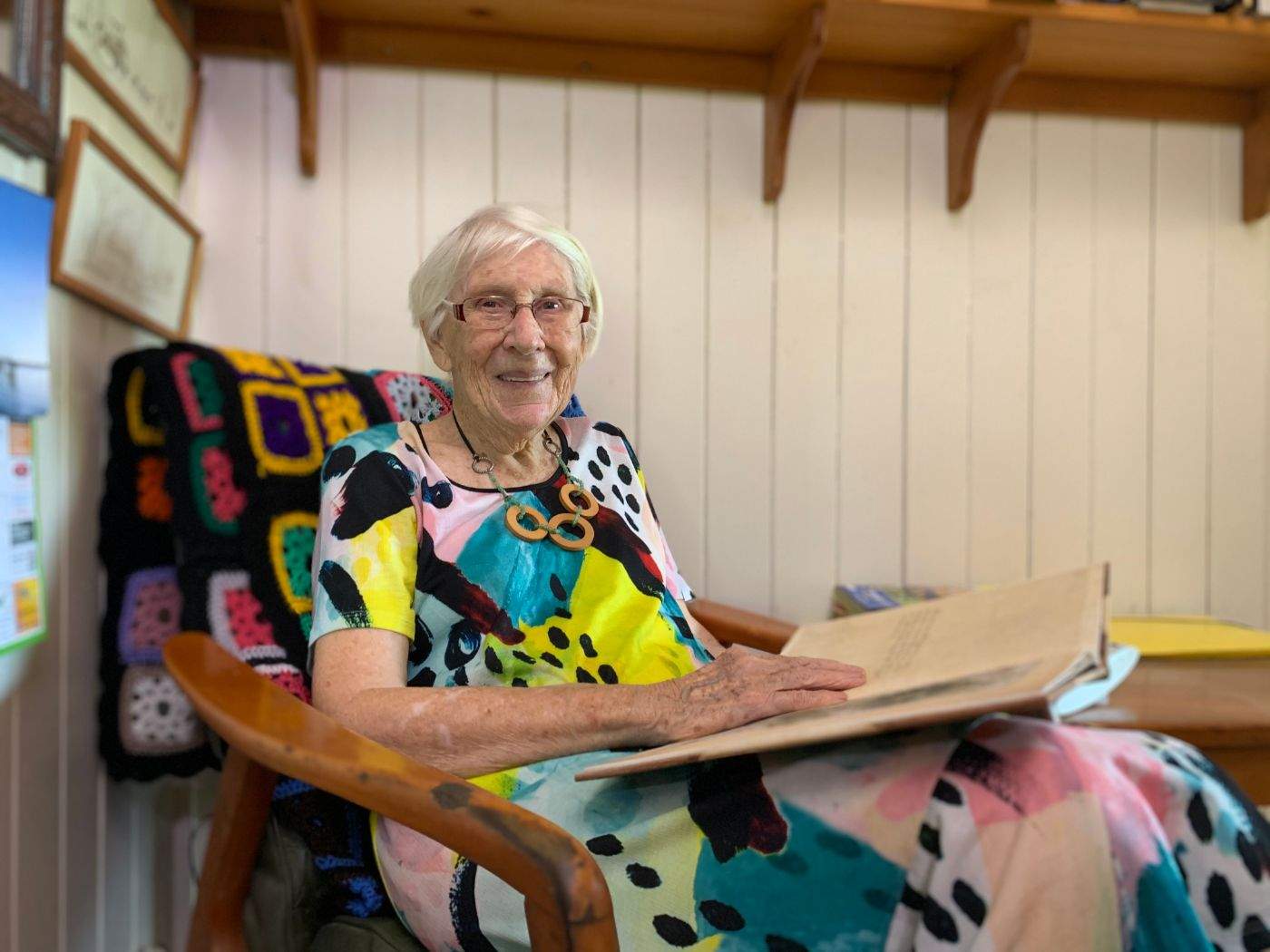 An elderly woman in a colourful dress sits in a rocking chair with a large book in her lap.