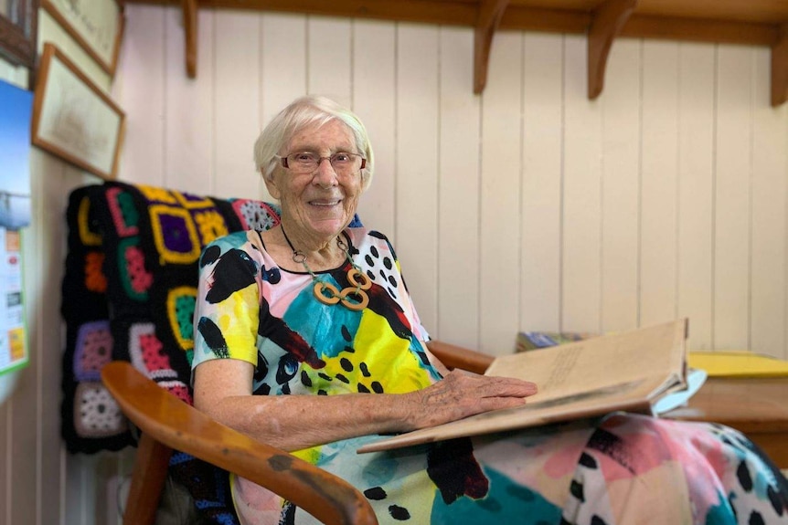 An elderly woman in a colourful dress sits in a rocking chair with a large book in her lap.