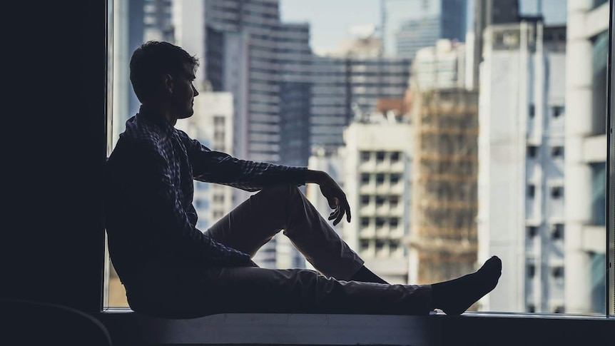 The silhouette of a man sitting on a windowsill, looking out over a city.