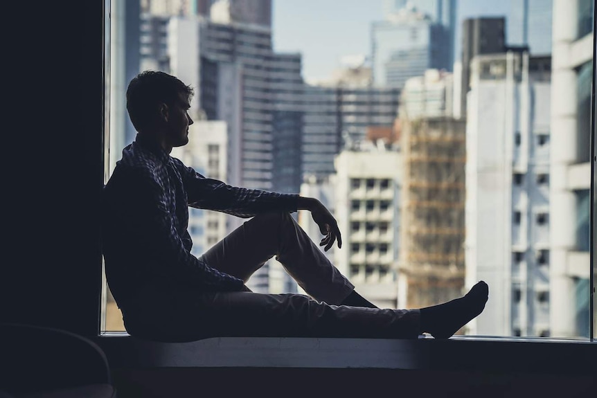 The silhouette of a man sitting on a windowsill, looking out over a city.