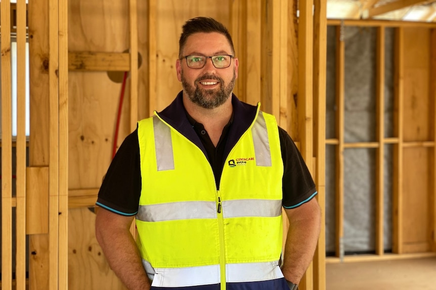 A bearded man in fluorescent tradie clothing and glasses stands in front of a construction site.