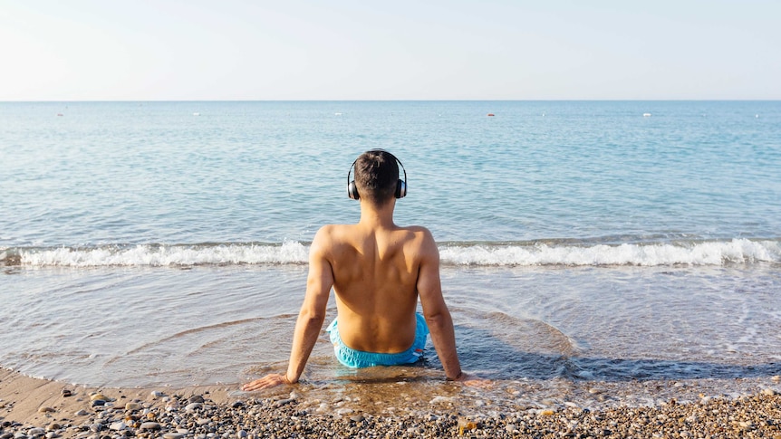 Rear view of a man sitting in the the water at the beach with headphones on.