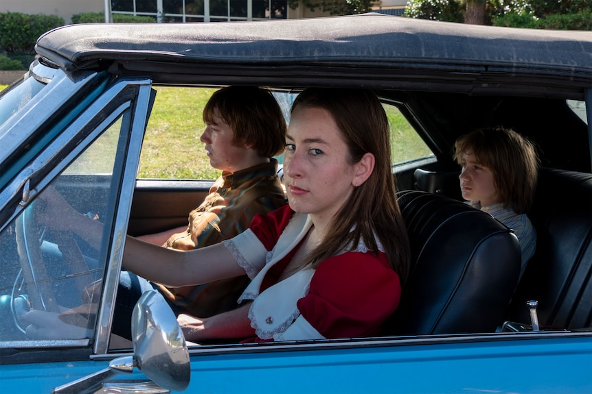 A teenage boy, a 20-something brunette woman and a young boy are driving in a blue car, each dressed in 70s-style clothing