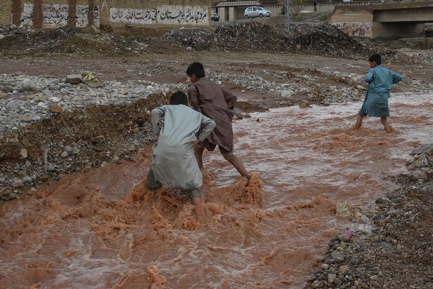 Children play in silty, brown, ankle-deep floodwater thats flowing rapildy near a damaged road.