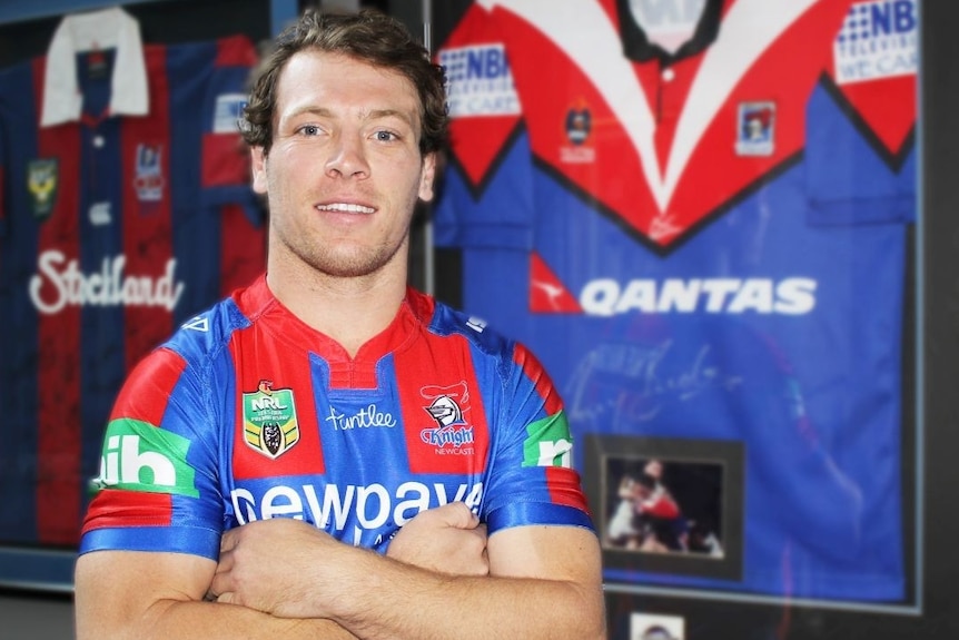 A man with short brown hair, wearing a Newcastle Knights jersey, standing in front of a framed jersey.