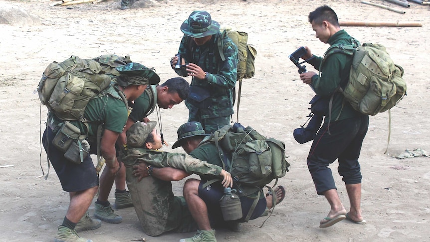 A group of men in camouflage gear, some holding video cameras, help a man sitting on the ground.