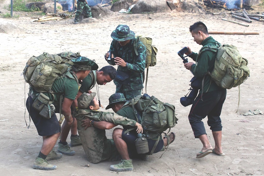 A group of men in camouflage gear, some holding video cameras, help a man sitting on the ground.