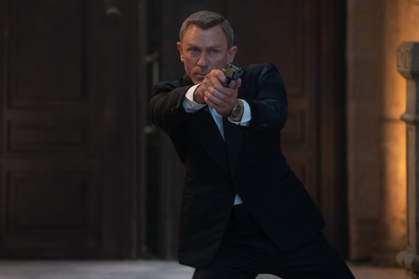 Middle-aged white man with fair cropped hair wears a black tuxedo and looks intensely while pointing a handgun