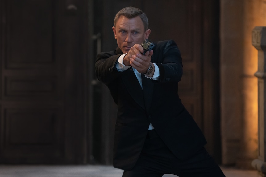 Middle-aged white man with fair cropped hair wears a black tuxedo and looks intensely while pointing a handgun