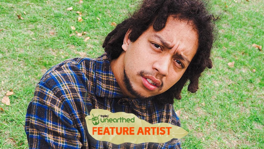The artist MALi JO$E sits on grass with his arms folded, looking at the camera.
