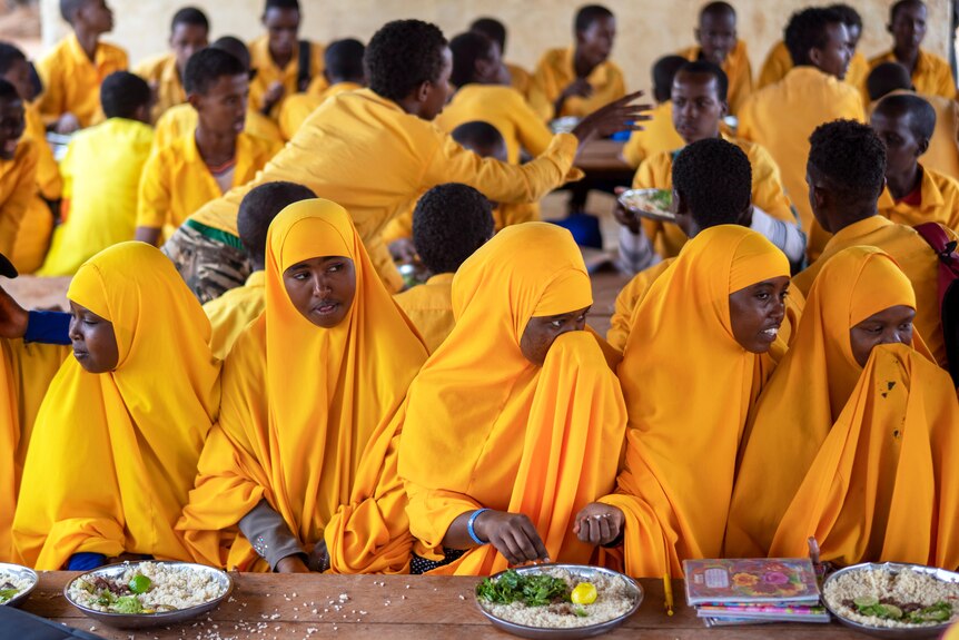 students dressed in yellow clothes sit and eat meals with rice at a school