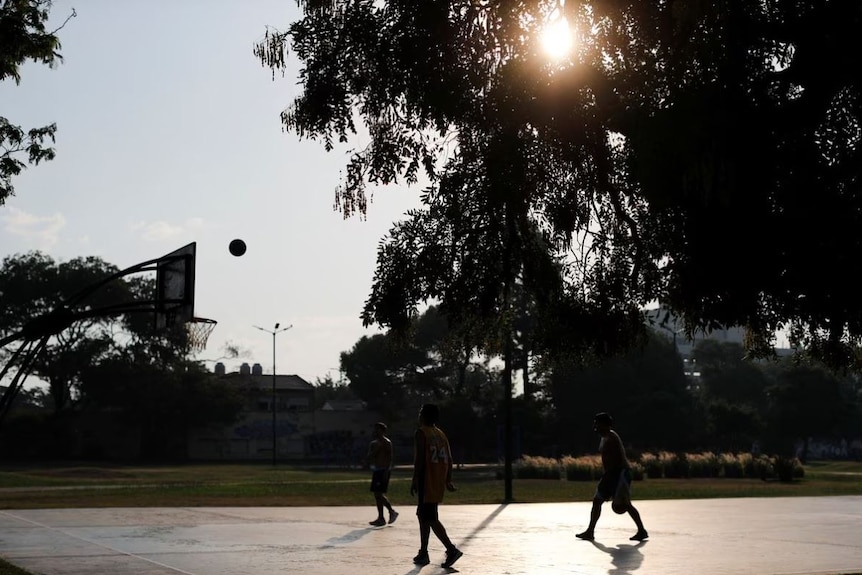 Three people play on a basketball court with the sun and trees in background