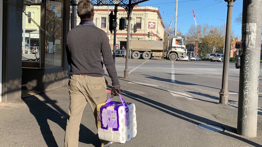 A man walks on the footpath holding a pack of toilet paper on a sunny day.