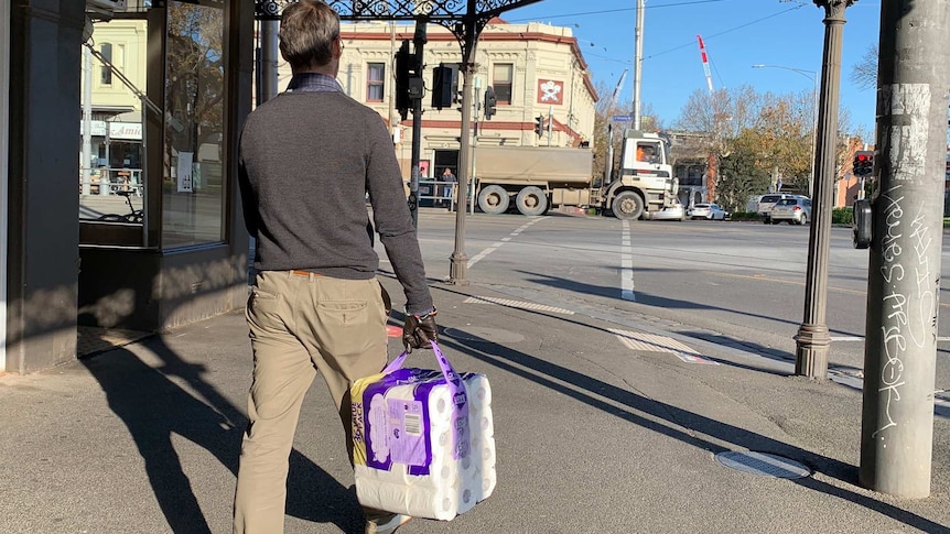 A man walks on the footpath holding a pack of toilet paper on a sunny day.