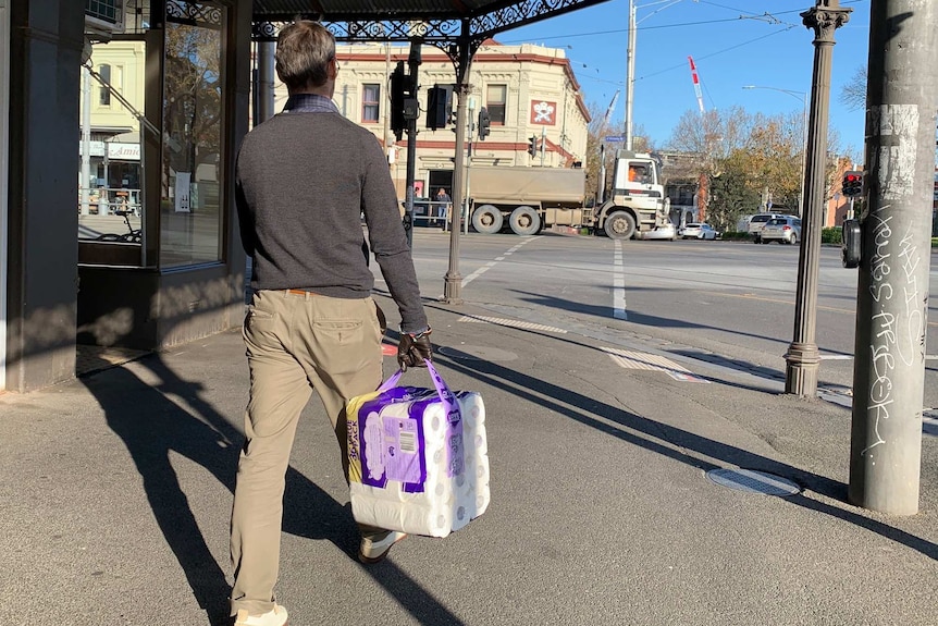 A man walks on the footpath holding a pack of toilet paper on a sunny day.