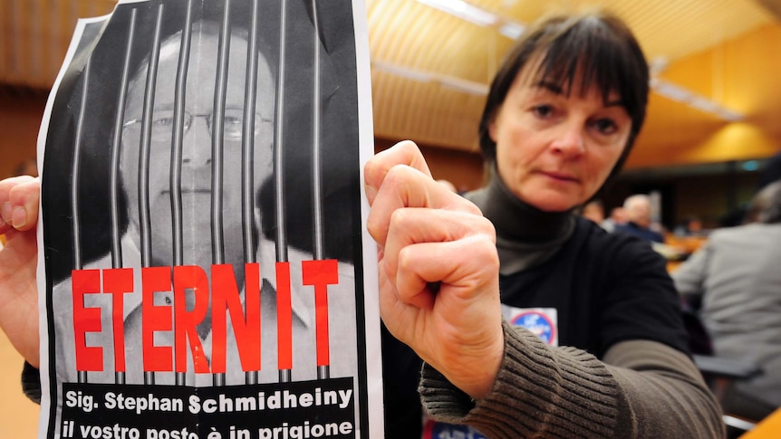 A woman holds a sign reading "Stephan Schmidheiny your place is in jail"