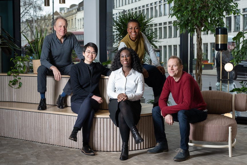 Two white men, two Black women and an Asian woman sit on stylish beige furniture, smiling.