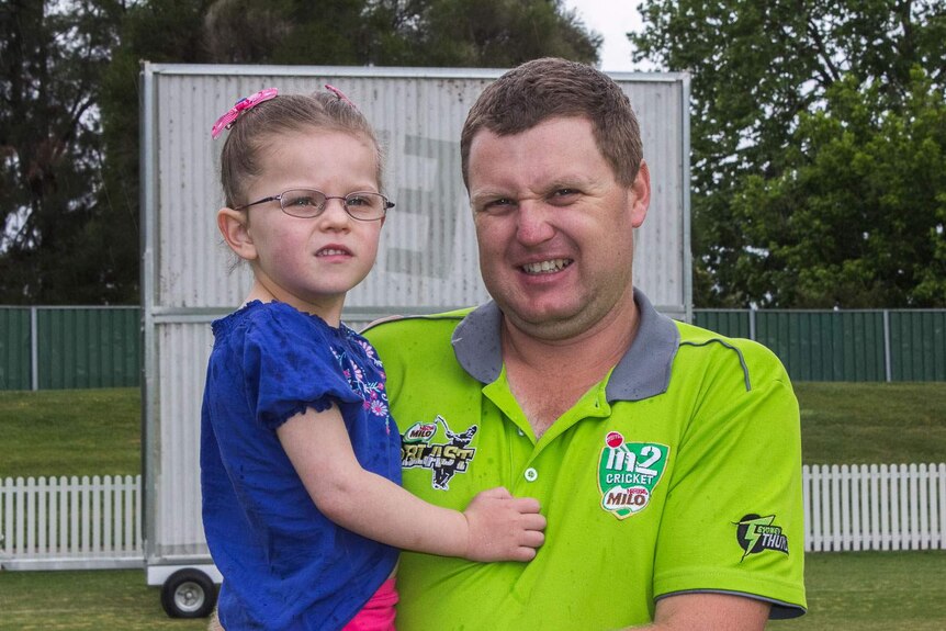 A cricket development manager in a green T-20 shirt holds his daughter, in the rain, in front of a cricket sight screen
