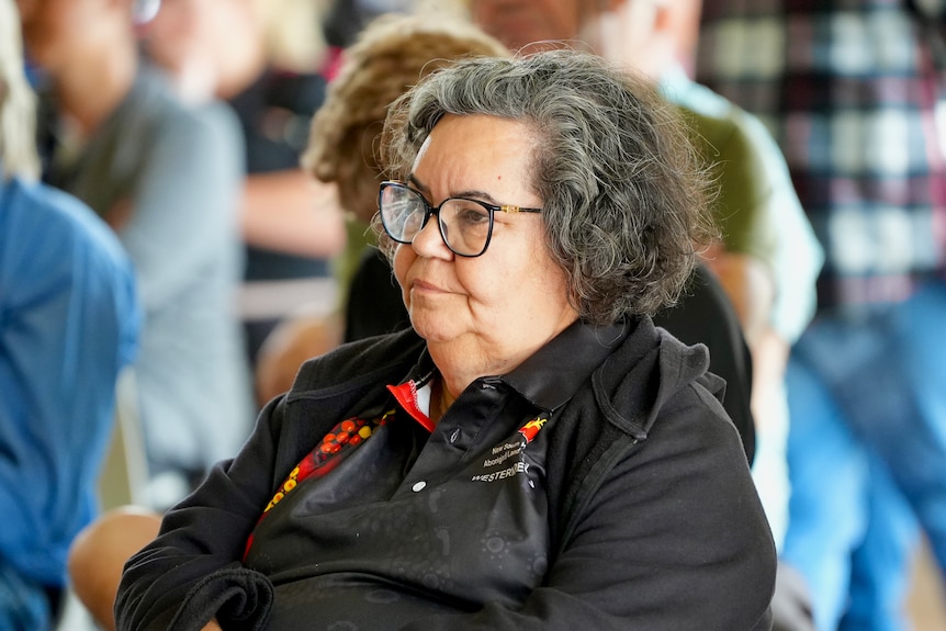 An older woman with greying hair sitting in a meeting, looking displeased.