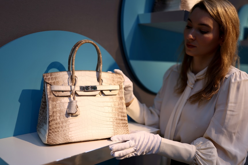 A woman in white gloves adjusts a Birkin bag on a shelf 