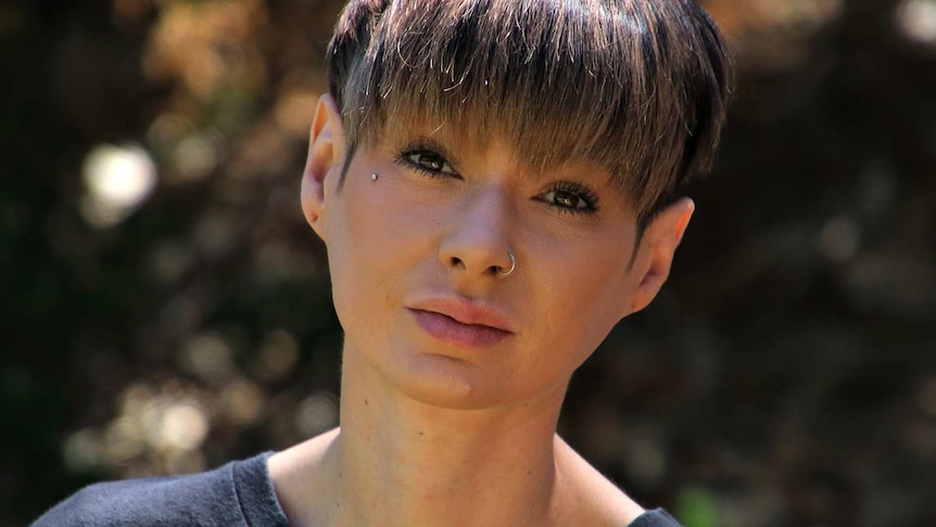 A tight head and shoulders shot of a woman with short hair wearing a black shirt and nose ring posing for a photo outdoors.