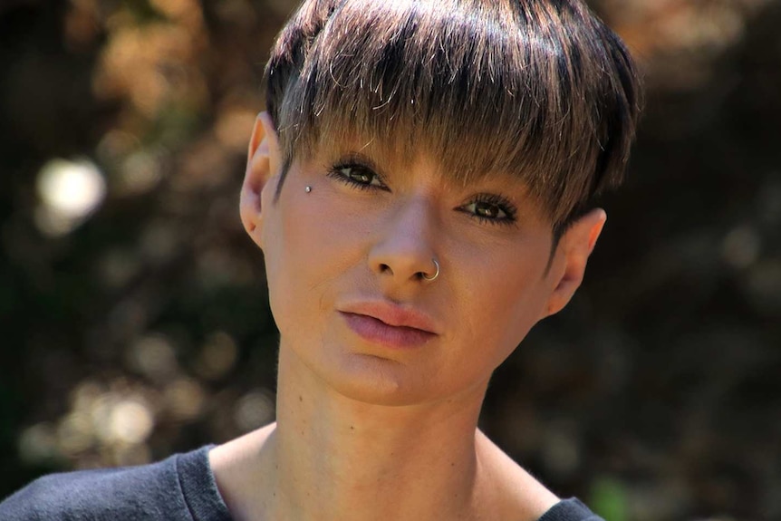 A tight head and shoulders shot of a woman with short hair wearing a black shirt and nose ring posing for a photo outdoors.