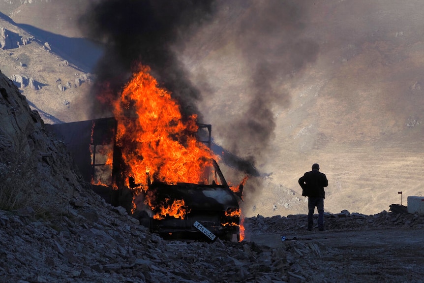 On a mountain road, you view a charred bus engulfed in flames near the black silhouette of a man looking down.