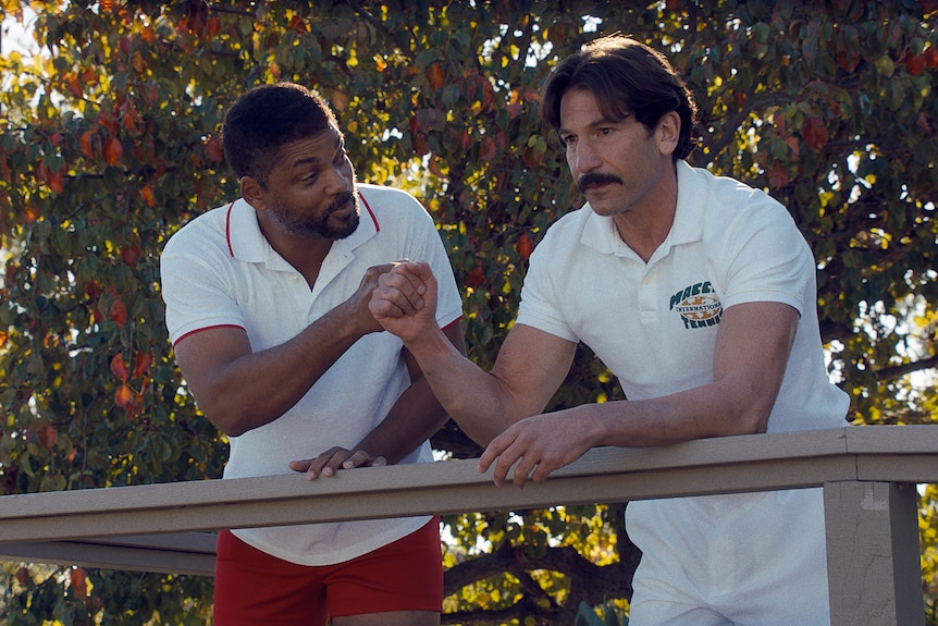 A middle-aged African American man leans over to speak intently to another sombre-looking man, each dressed in tennis clothing