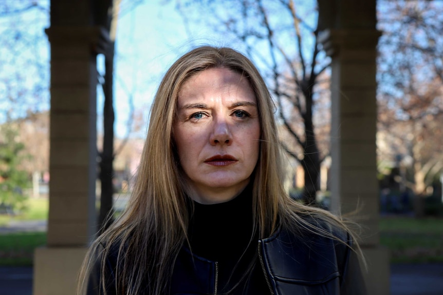 Woman with blonde hair and red lipstick stands beneath stone archway with dappled light across face