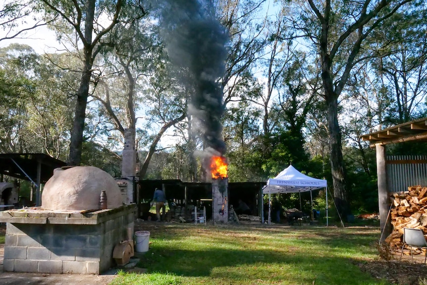 A wood burning kiln sends smoke into a tree laden enclave. 