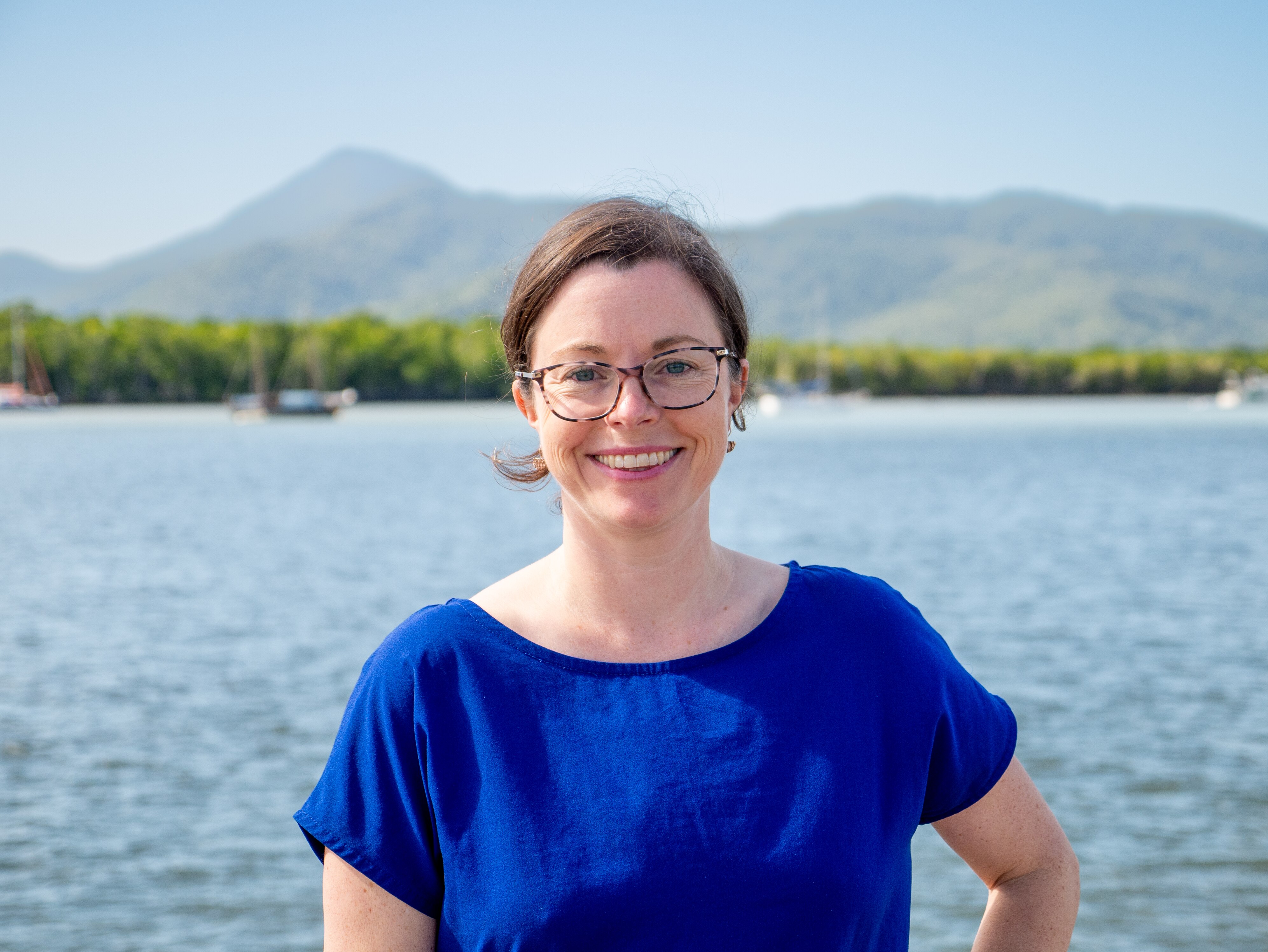 A portrait image of a smiling woman wearing glasses and a blue top, standing in front of the sea and mountains in the background.