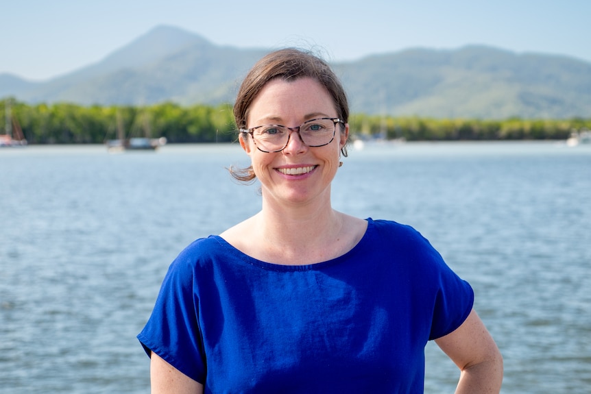 A portrait image of a smiling woman wearing glasses and a blue top, standing in front of the sea and mountains in the background.