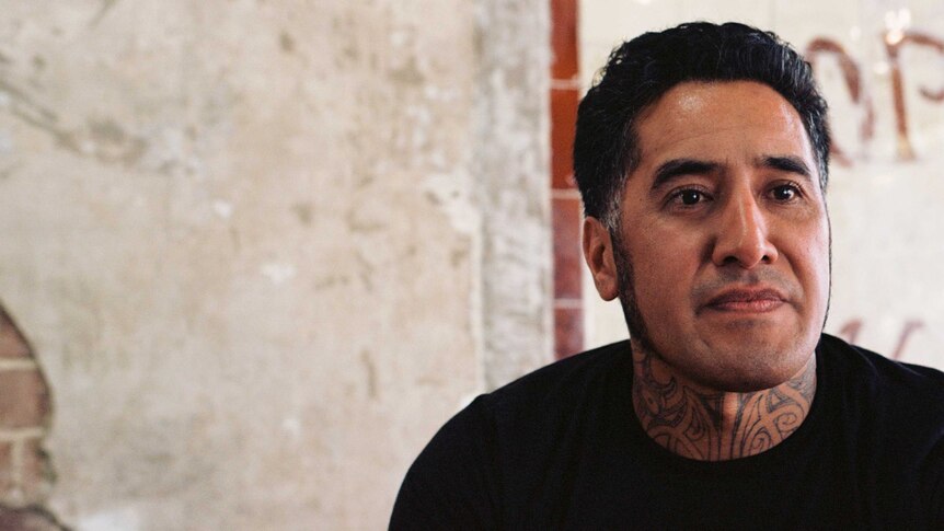 A portrait of a Maori man in a black T-shirt, gazing slightly to the side of the camera. A brick wall is seen in the background.