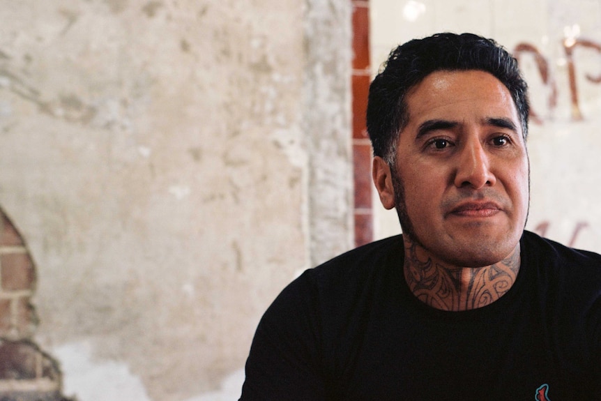 A portrait of a Maori man in a black T-shirt, gazing slightly to the side of the camera. A brick wall is seen in the background.