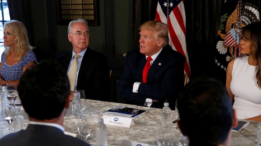 Donald Trumps speaks with his arms folded while sitting at a table flanked by Melania Trump and Tom Price.