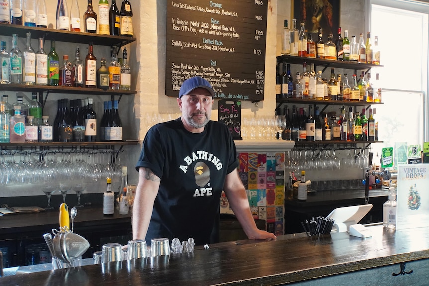A man wears a baseball cap and black T-Shirt while standing behind a bar