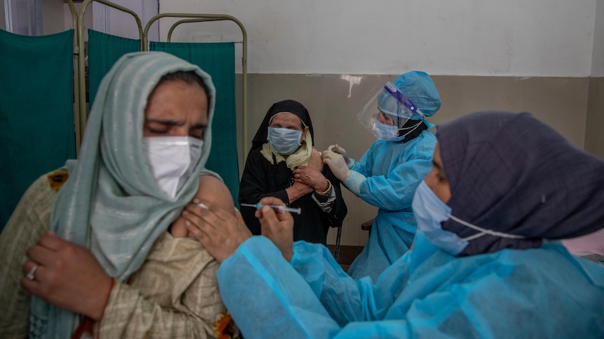Nurses wearing blue gowns and face shields inject women wearing headscarves at a health clinic.