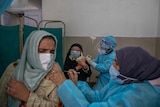 Nurses wearing blue gowns and face shields inject women wearing headscarves at a health clinic.