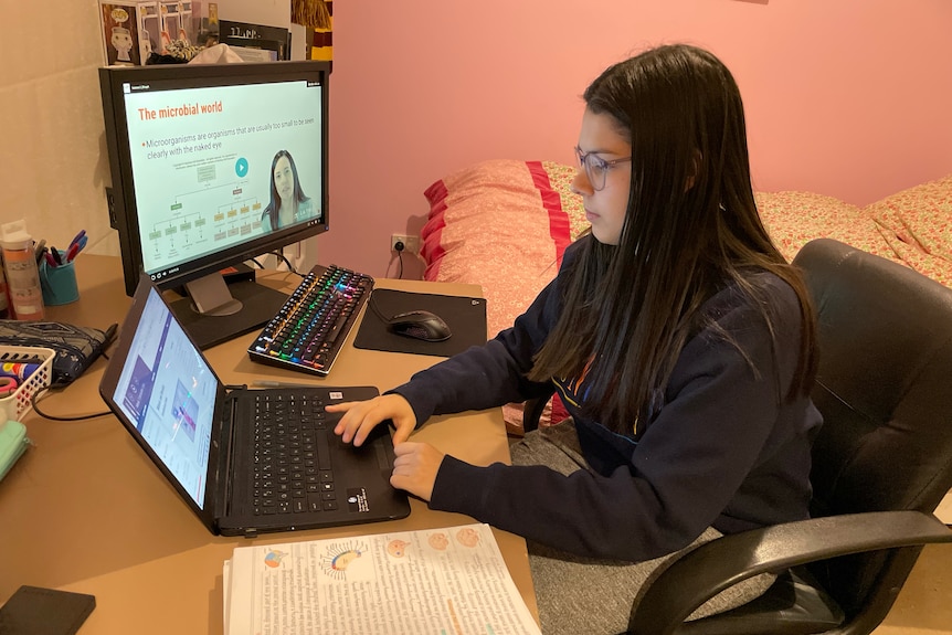 A woman with glasses is sitting at her computer in her bedroom