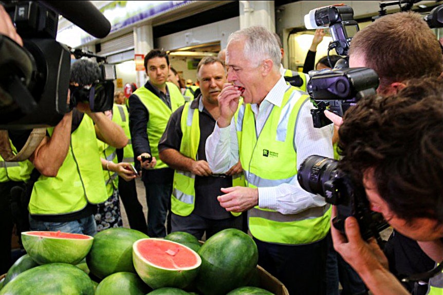 Side on view of Malcolm Turnbull eating some watermelon with a large crate of watermelons in the foreground.