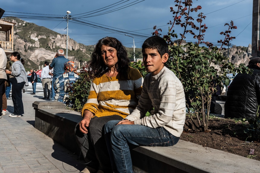 A woman and her son sit on a bench near a tree in the sun.