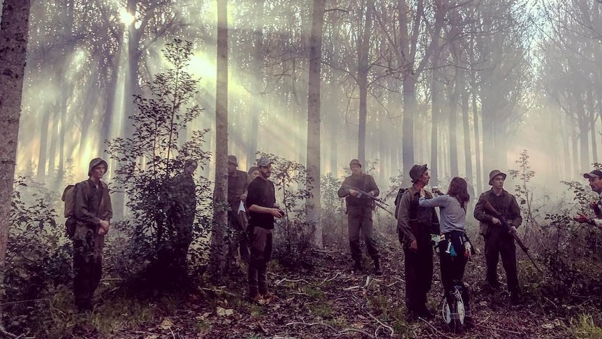 People dressed as soldiers and carrying guns stand beneath a forest of tall trees as the sun's rays pierce through the trees.