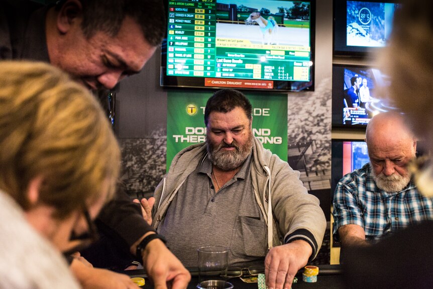 Ken Skinner sitting at a poker table surrounded by five people with a  television screen above his head.