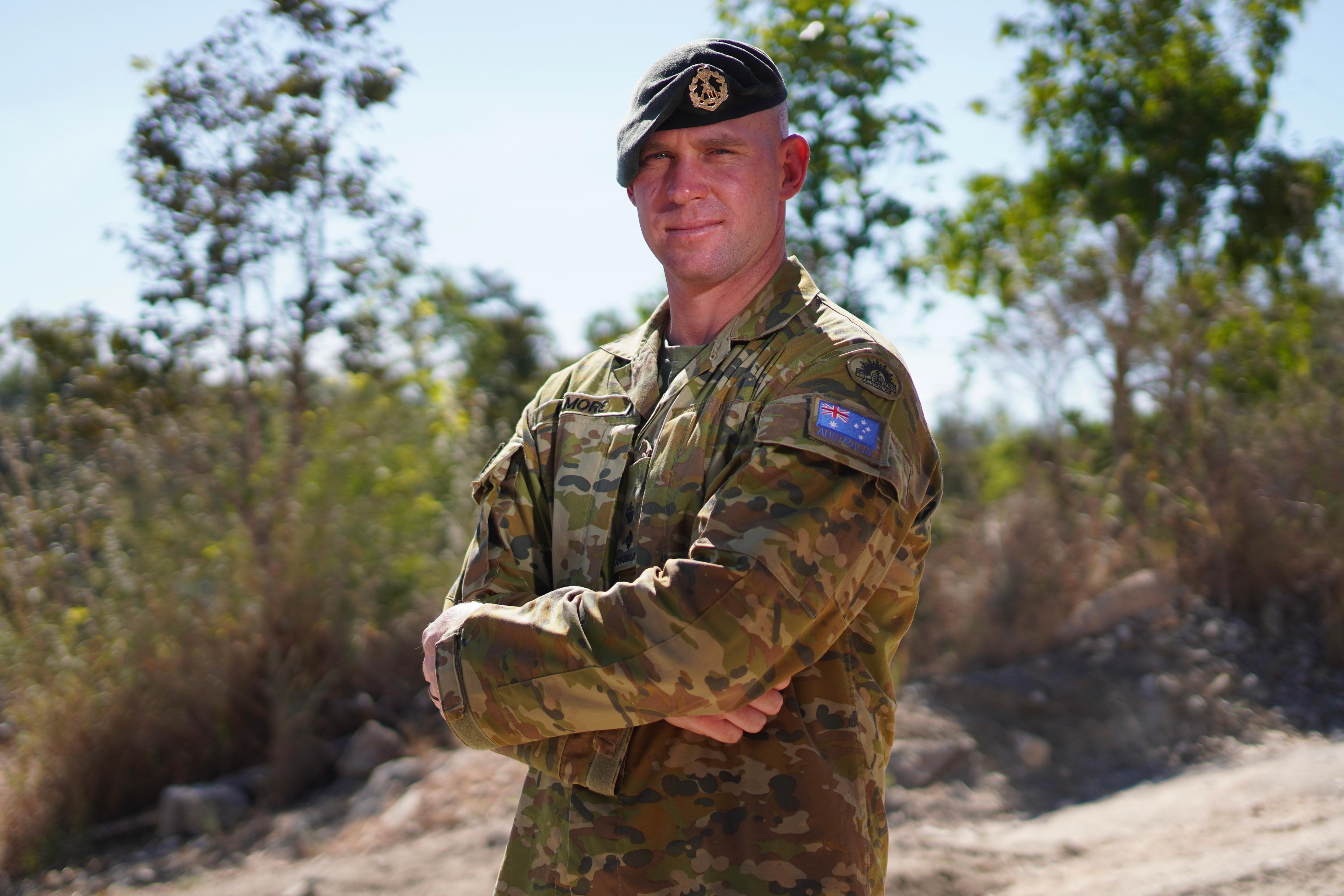 A man wearing a green army hat and a khaki army outfit looks directly at the camera with his arms crossed.